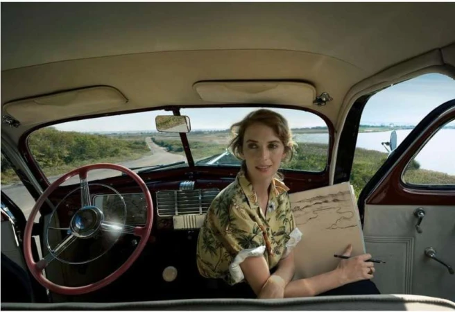 Woman inside classic car drawing landscape. Female adult with sketchpad in vintage vehicle against road and distant water view.