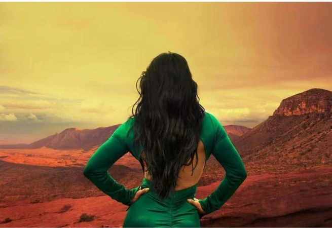 Woman with long dark hair seen from behind wearing a green dress against a desert mountain landscape at sunset