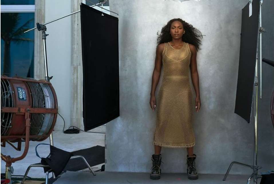 Full-length portrait of a Black woman in a sheer gold dress and black boots posing in a photo studio with professional lighting equipment.