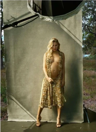 Pregnant blond woman standing in front of a grey seamless backdrop wearing an open patterned dress and heels.