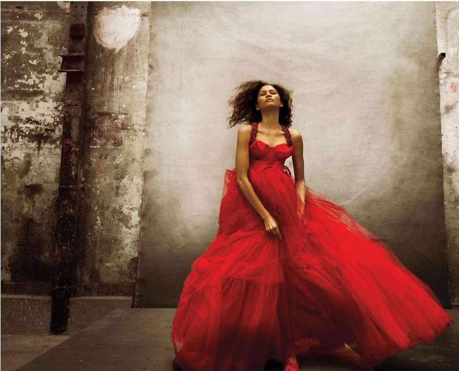 A woman in a long flowing red dress standing in a dilapidated interior with concrete walls and floors and overhead lighting.