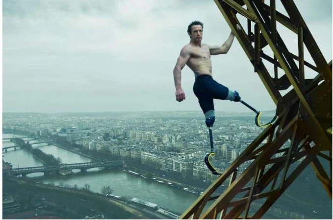 A disabled man with prosthetic legs climbs a metal structure, with a cityscape and overcast sky as a backdrop, action shot.