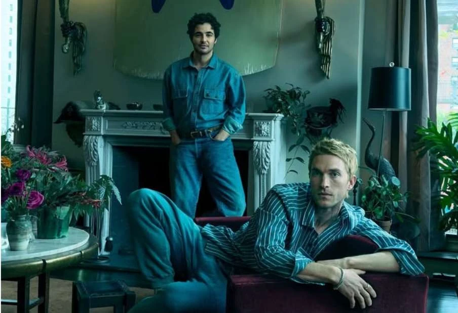 Two men pose indoors in casual denim and striped outfits. Interior includes plants, decor, natural light, creating an intimate setting.