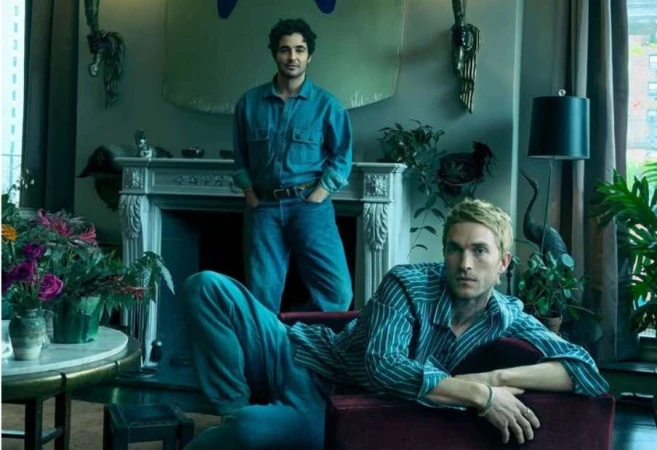 Two men pose indoors in casual denim and striped outfits. Interior includes plants, decor, natural light, creating an intimate setting.