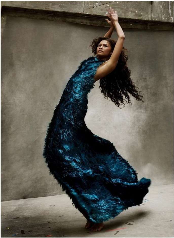 Zendaya posing in a textured dark blue gown with arms raised against a neutral concrete background