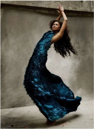 Zendaya posing in a textured dark blue gown with arms raised against a neutral concrete background