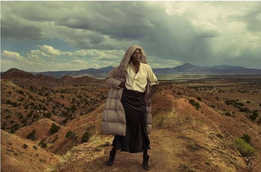 An adult female fashion model poses outdoors in a puffer coat and black skirt, standing on a desert ridge with a cloudy sky backdrop.