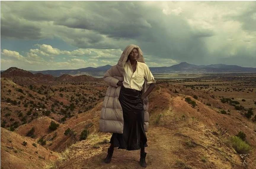 An adult female fashion model poses outdoors in a puffer coat and black skirt, standing on a desert ridge with a cloudy sky backdrop.