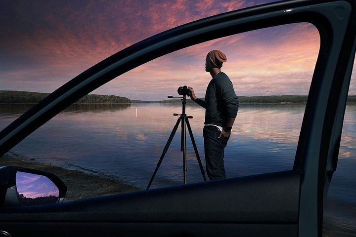 Man photographs lake sunset with tripod-mounted camera, viewed through car window. Calm water and sky reflect pastel light.