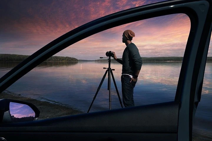 Man photographs lake sunset with tripod-mounted camera, viewed through car window. Calm water and sky reflect pastel light.