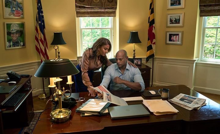Two adults at a large wooden desk indoors, reading a document, with American and Maryland flags visible. Natural light illuminates the room.