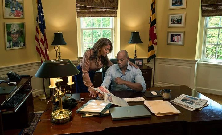 Two adults at a large wooden desk indoors, reading a document, with American and Maryland flags visible. Natural light illuminates the room.