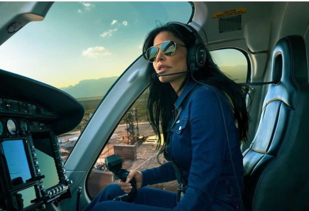 Woman in a helicopter cockpit with sunglasses and headset, holding controls; arid landscape visible in the background through the window.