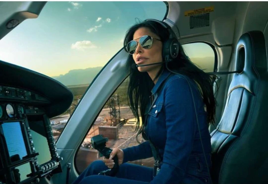 Woman in a helicopter cockpit with sunglasses and headset, holding controls; arid landscape visible in the background through the window.