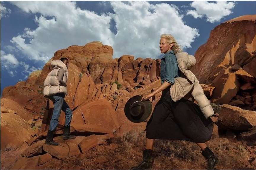 Two people in outdoor fashion shoot. One woman walks with a hat and jacket, another stands on rocks, set against rock formations and sky.