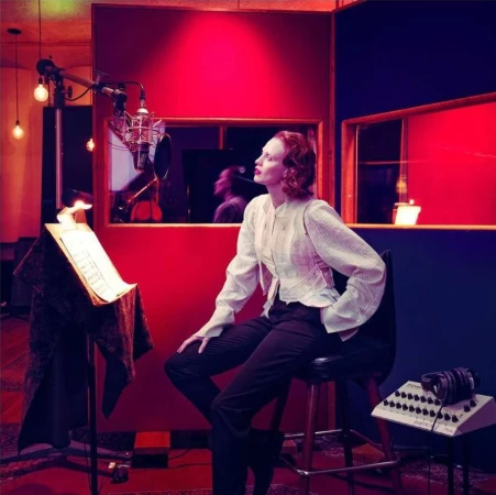 Woman in striped top and trousers poses in a vintage-style recording studio with a microphone, music stand, and sound equipment.