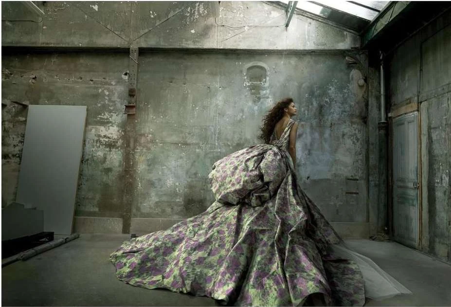 Woman in floral gown poses in abandoned building with peeling paint, arched windows, and concrete floors in a high-fashion editorial shoot.