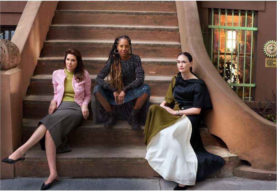 Three women are posing on a stoop with an urban background. They are wearing stylish, fashionable clothing. The location is a city street.