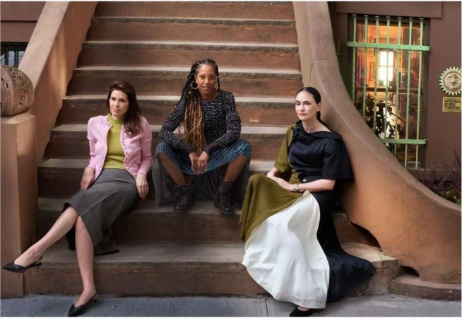 Three women are posing on a stoop with an urban background. They are wearing stylish, fashionable clothing. The location is a city street.