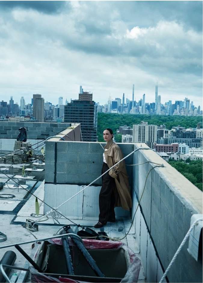 A woman wearing a long tan trench coat standing on a concrete rooftop overlooking a city skyline under a cloudy sky.