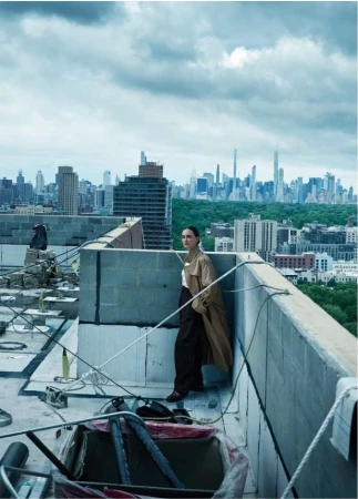 A woman wearing a long tan trench coat standing on a concrete rooftop overlooking a city skyline under a cloudy sky.