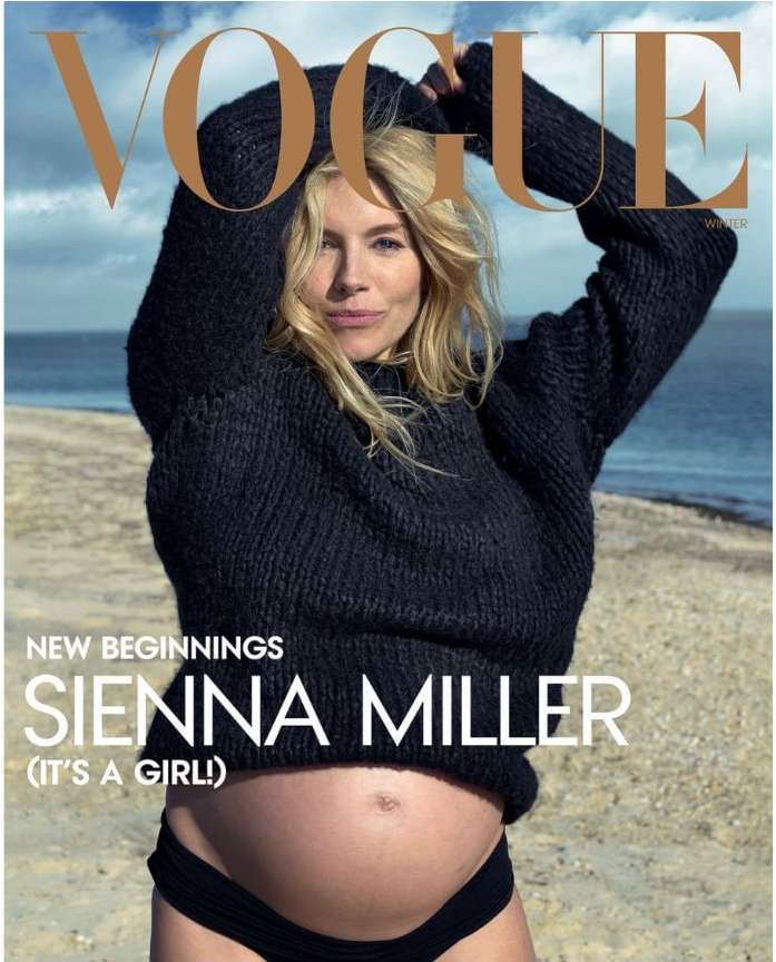 Sienna Miller poses on a beach for Vogue magazine cover. The pregnant woman wears a knit sweater and swimsuit bottoms in a sunny outdoor setting.