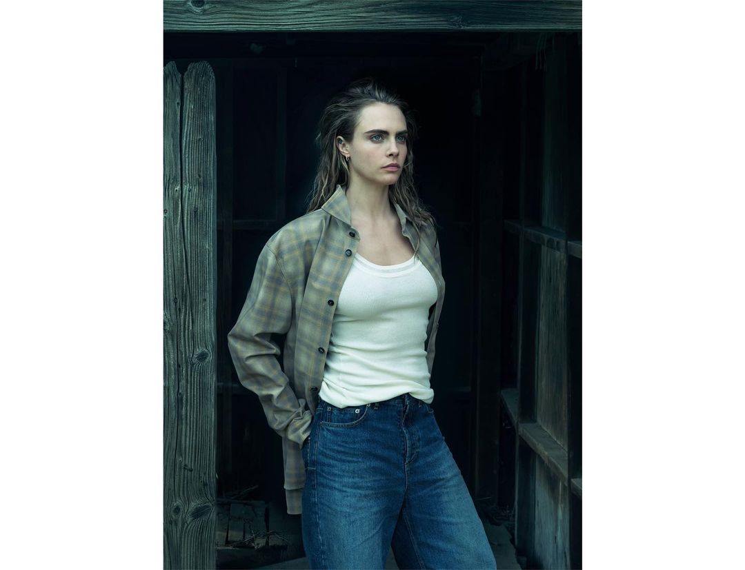 Cara Delevingne posing in a plaid shirt and blue jeans in front of a dark rustic wooden structure.