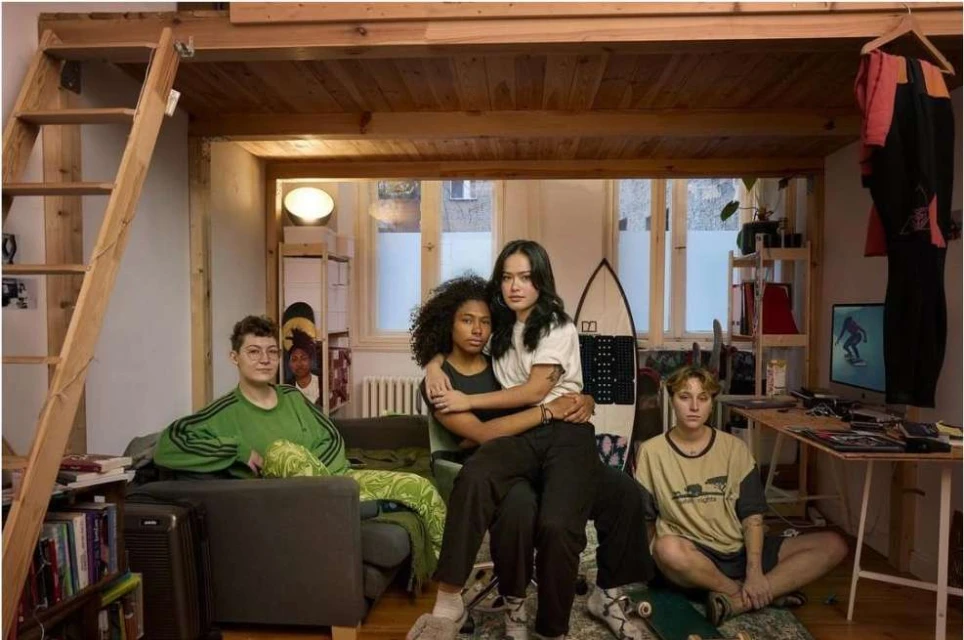 Group portrait in a rustic bedroom shows four young adults sitting and relaxing near surfboards, skateboards, and various personal items.
