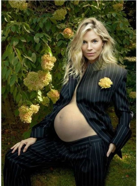 A pregnant woman with blonde hair poses in a pinstripe suit outdoors, with yellow flowers and green foliage in the background.