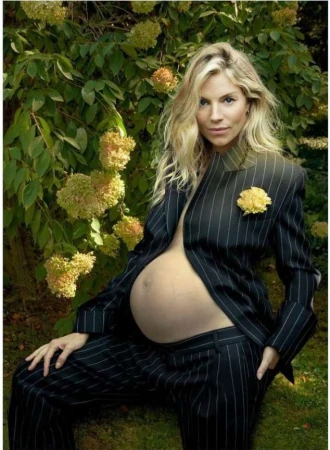 A pregnant woman with blonde hair poses in a pinstripe suit outdoors, with yellow flowers and green foliage in the background.