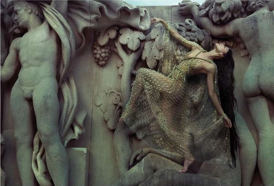 Elegant woman in a beaded dress gracefully poses among detailed stone statues, showcasing a blend of fashion and classical art.