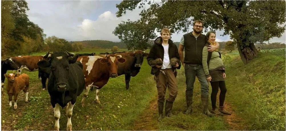 Family portrait featuring a mother, father, and child, along with a group of cows in a rural, green pasture setting with trees.
