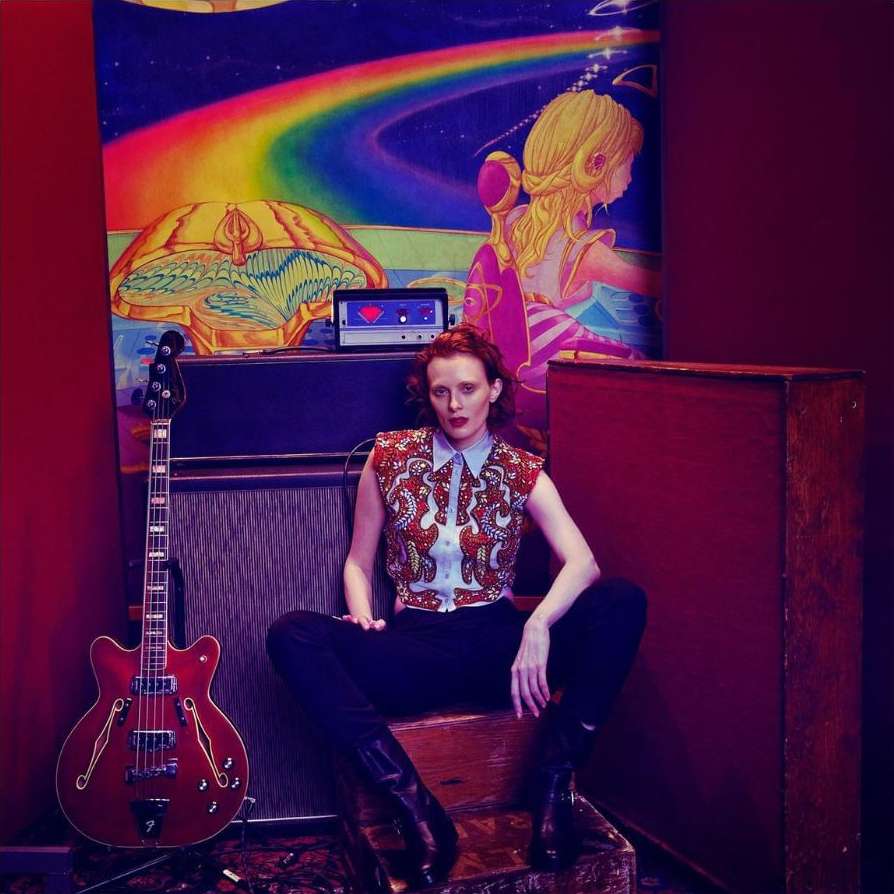 Stylish woman in a patterned top poses indoors with a cherry red guitar against a colorful wall tapestry and vintage equipment.