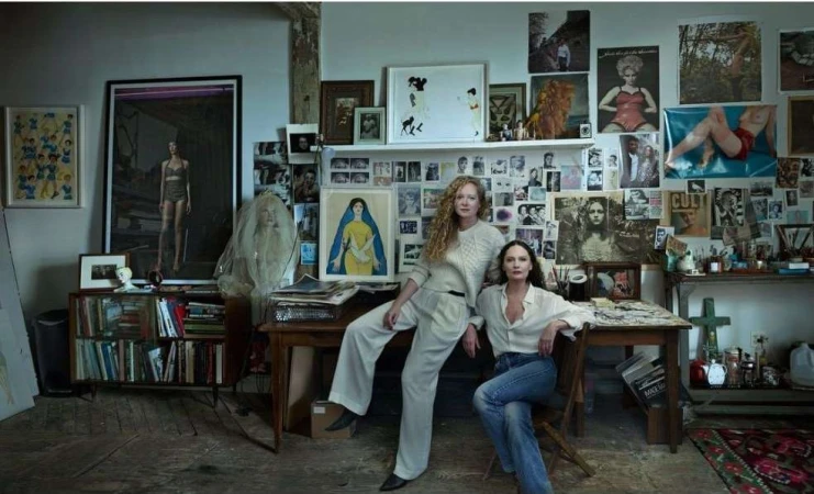 Two women pose in an artist's studio filled with artwork, posters, books, and creative clutter, creating an intimate and artistic scene.