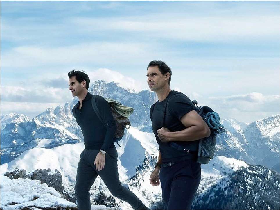 Two adult males wearing backpacks climb a snow-covered mountain with a backdrop of distant snow-capped peaks under a bright sky.
