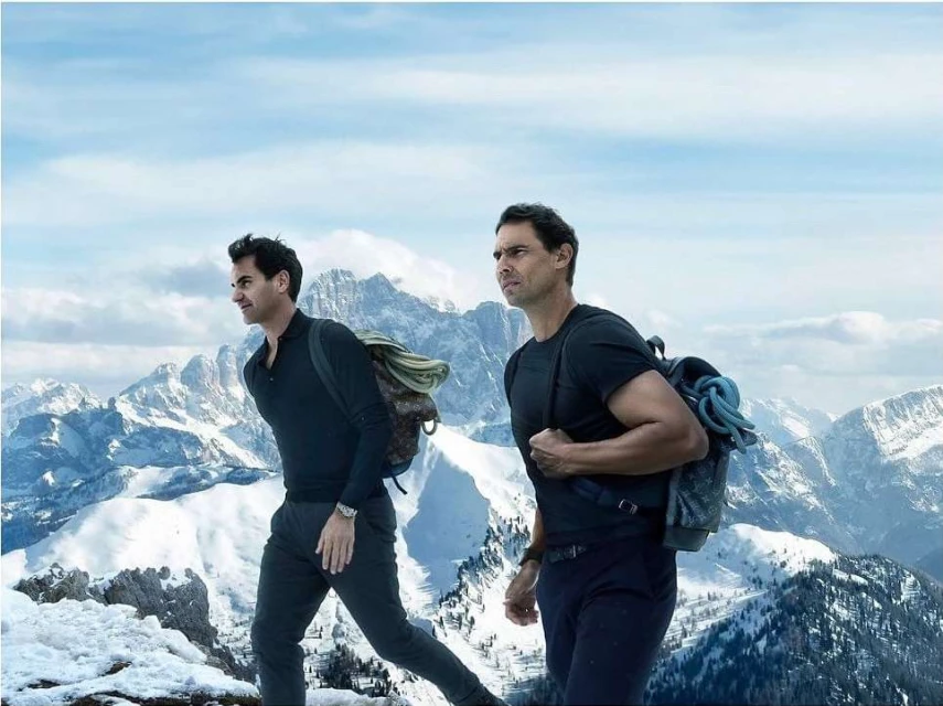 Two adult males wearing backpacks climb a snow-covered mountain with a backdrop of distant snow-capped peaks under a bright sky.