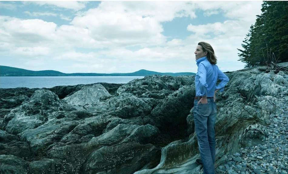 Adult female standing on a rocky beach looking out at the ocean on a cloudy day; trees and mountains in the distance.
