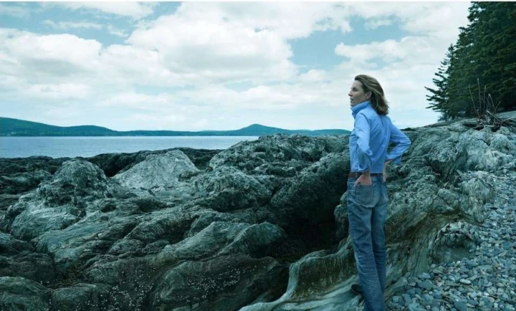 Adult female standing on a rocky beach looking out at the ocean on a cloudy day; trees and mountains in the distance.