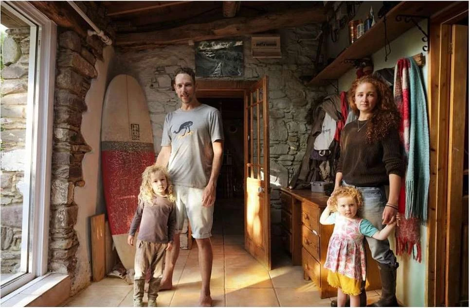 Family of four poses inside a rustic home with stone walls. A surfboard stands to one side, adding a touch of coastal style to the interior.