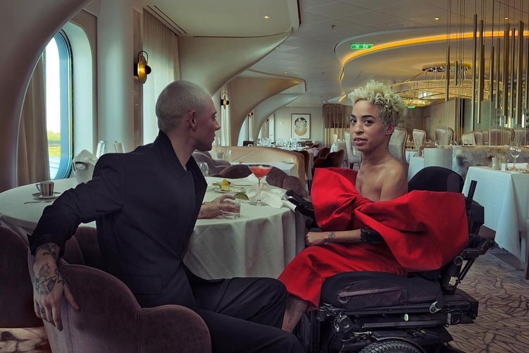 Couple seated at a restaurant table, one in wheelchair wearing a large red bow, dining indoors with modern interior design.