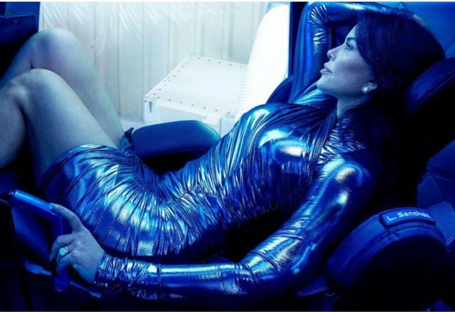 Adult woman reclines in a futuristic chair wearing a shiny blue dress. Blue lighting dominates the scene. The setting appears to be indoors.