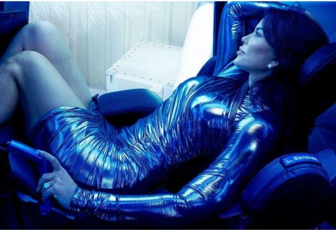 Adult woman reclines in a futuristic chair wearing a shiny blue dress. Blue lighting dominates the scene. The setting appears to be indoors.