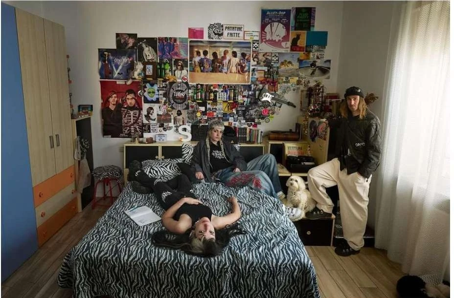 Three young adults relaxing in a bedroom decorated with posters and zebra-print patterns, featuring a small white dog on the bed.