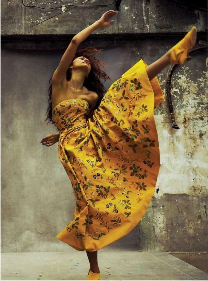 Woman in a patterned yellow strapless dress dances with one leg extended upwards against a textured grey wall in a full shot.