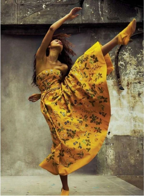 Woman in a patterned yellow strapless dress dances with one leg extended upwards against a textured grey wall in a full shot.