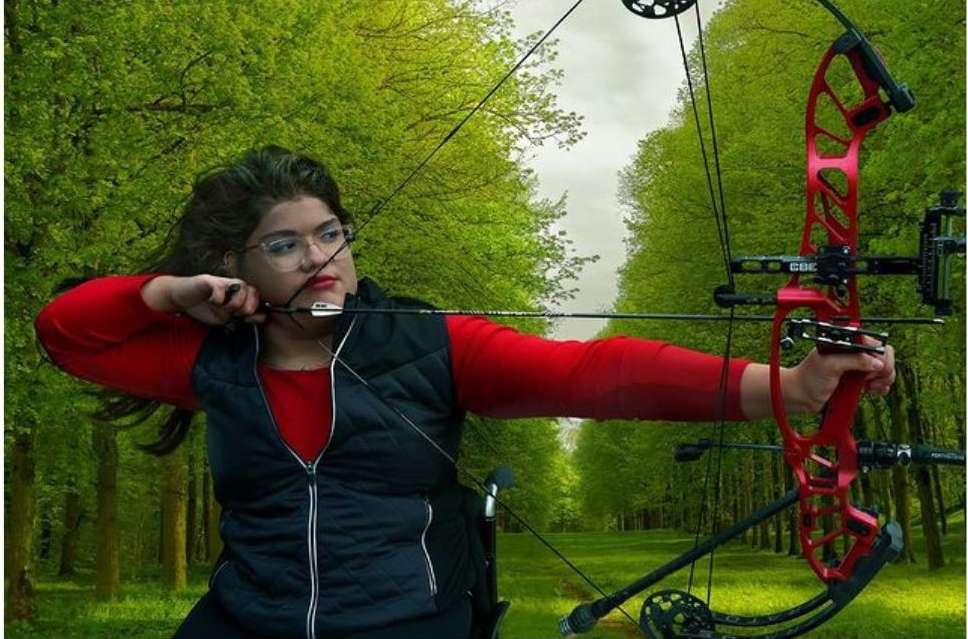 Adult female archer with glasses in outdoor forest setting practices archery with red compound bow and arrow, wearing red shirt and black vest.