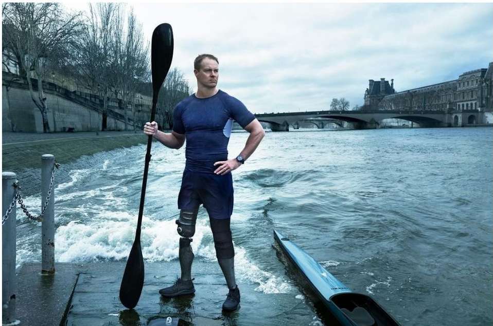 Adult male athlete with prosthetic leg stands on a quay with paddle, with river, bridge, and buildings in the background on a cloudy day.