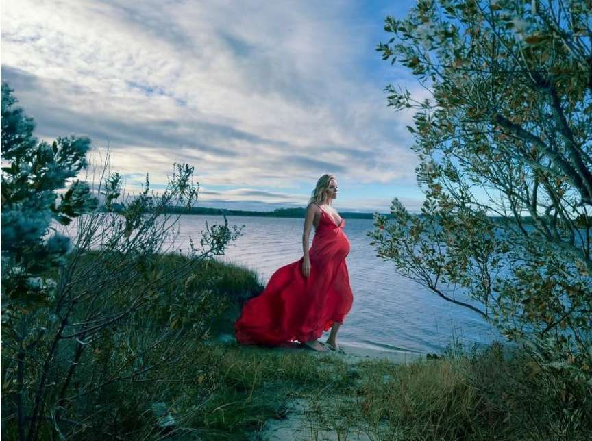 Pregnant woman in a flowing red dress stands near the ocean surrounded by trees under a partly cloudy sky in a serene outdoor setting.