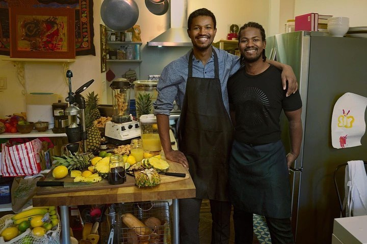 Two smiling men stand indoors with fresh fruit and a blender. The kitchen features pineapples, lemons, and limes.