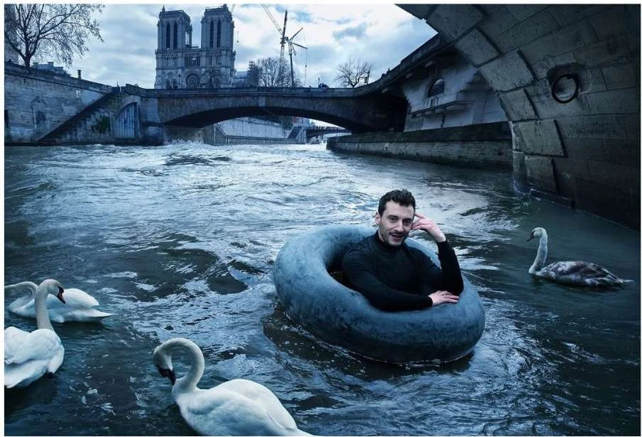 Adult male sits in an inner tube floating on a river with swans, in front of a bridge and the Notre Dame Cathedral in Paris, France.